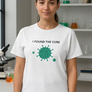 Può includere: T-shirt bianca con la scritta "I FOUND THE CURE" e un disegno di un virus verde. La persona indossa la maglietta in un ambiente di laboratorio, con attrezzature scientifiche visibili sugli scaffali sullo sfondo.