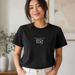 Puede incluir: Camiseta negra de cuello redondo con el texto "Just one more tile" y un gráfico de dos fichas. La camiseta se combina con pantalones blancos. La imagen está tomada en interiores.