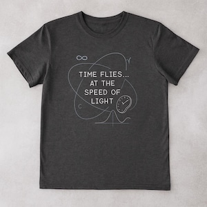 Può includere: T-shirt grigio scuro con la scritta bianca "TIME FLIES... AT THE SPEED OF LIGHT". Il design include un simbolo di infinito, un orologio e grafici scientifici. Una t-shirt casual e unisex.