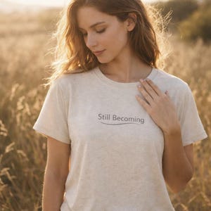 Può includere: Una maglietta beige chiaro con la scritta "Still Becoming" in un carattere grigio scuro. La maglietta ha maniche corte e girocollo. La modella indossa la maglietta in un campo di erba alta.