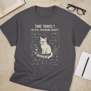 May include: Dark gray t-shirt featuring a white cat wearing glasses, sitting on a book, with scientific equations and the text "TIME TRAVEL? I'M STILL DEBUGGING GRAVITY." A fun, science-themed graphic tee.