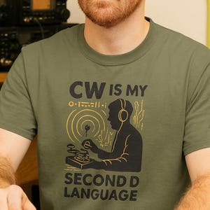Può includere: T-shirt verde oliva con la scritta "CW IS MY SECOND LANGUAGE". Il design presenta la silhouette di una persona con le cuffie, che utilizza un tasto telegrafico, con un disegno di onde radio dorate. Girocollo.