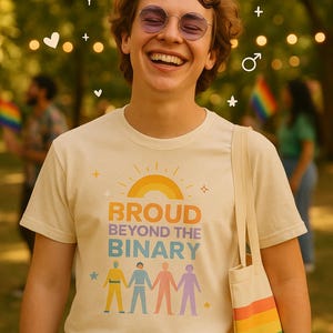 Può includere: Una persona indossa una maglietta color crema con la scritta "PROUD BEYOND THE BINARY" e un arcobaleno. La persona sorride e tiene una borsa con strisce arcobaleno.