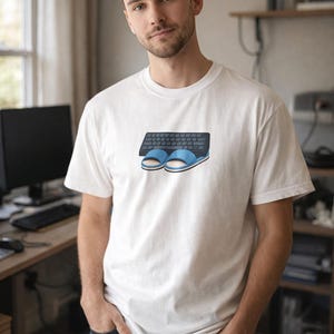 Può includere: T-shirt bianca con grafica di tastiera nera e pantofole blu. La maglietta è realizzata in materiale morbido. La persona indossa jeans blu scuro.