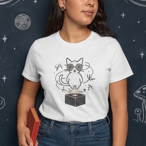 May include: White t-shirt featuring a cartoon cat wearing glasses and standing on a black cube. The cat is surrounded by scientific symbols. The background is a dark blue chalkboard with drawings of planets and stars.
