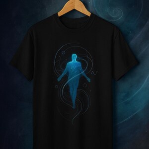 May include: Black t-shirt featuring a blue silhouette of a person with outstretched arms, surrounded by swirling lines and dots. The design has a cosmic or ethereal theme, suggesting a sense of space or spirituality. The shirt is hanging on a wooden hanger.