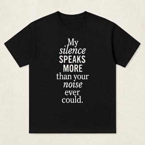 Può includere: T-shirt nera con la scritta bianca "My silence SPEAKS MORE than your noise ever could." La maglietta ha maniche corte e girocollo. Il testo è in un carattere stilizzato.