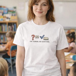 Può includere: T-shirt bianca con una grafica raffigurante un punto interrogativo rosso, un segno di spunta blu e un libro aperto. Il testo "TINY HUMANS. BIG QUESTIONS." è sotto la grafica. La maglietta è indossata da una persona in classe.