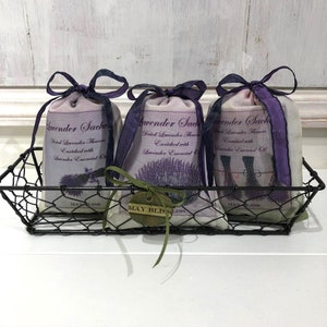 May include: Three lavender sachets in a wire basket. Each sachet is made of white fabric with a purple ribbon and a label that reads "Lavender Sachet" with the text "Dried Lavender Flowers Enriched with Lavender Essential Oil".