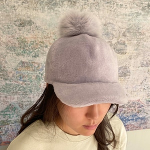 May include: A light purple fuzzy baseball cap with a faux fur pom pom on top.