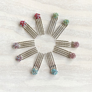 May include: Ten silver hair clips with colourful jeweled balls on the ends. The clips are arranged in a circle on a light wood background.