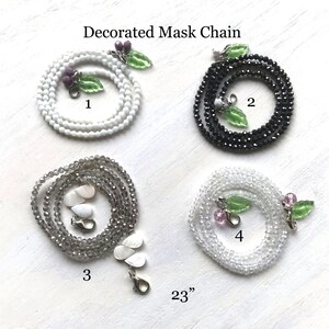 May include: Four different styles of decorated mask chains, each 23 inches long. The chains are made of white, black, gray, and clear beads. Each chain has a different charm attached to it, including a leaf with purple berries, a leaf with white berries, a shell, and a leaf with pink berries.
