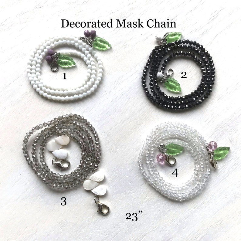 May include: Four different styles of decorated mask chains, each 23 inches long. The chains are made of white, black, gray, and clear beads. Each chain has a different charm attached to it, including a leaf with purple berries, a leaf with white berries, a shell, and a leaf with pink berries.