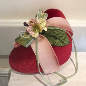 Op de afbeelding: Een rood fluwelen hartvormig kussen met een groen blad en een roze en groene strik met een bloemmotief.