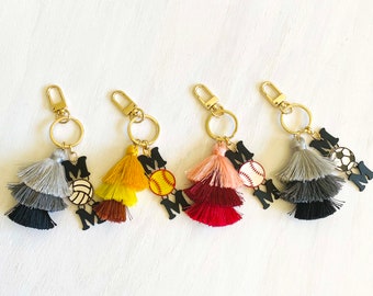 May include: Four keychains with colorful tassels and black letter charms that spell "MOM". Each keychain features a different sports ball charm: volleyball, softball, baseball, and soccer.