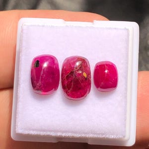 May include: Three polished, oval-shaped, deep red gemstones are displayed in a white plastic case. The gemstones vary slightly in size and have inclusions and imperfections. The gems are a vibrant, saturated colour.