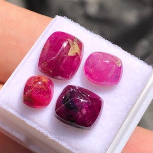 May include: Four polished, cushion-cut gemstones in varying shades of red and pink are displayed on a white surface. The gems have a smooth, rounded shape and exhibit a natural, translucent quality. The gemstones are likely rubies.