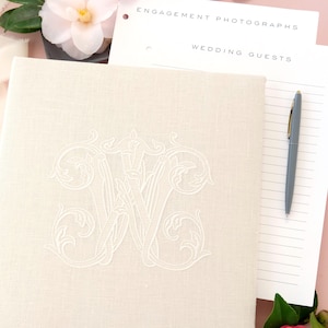 May include: A white linen wedding album with a white embroidered monogram of the letters 'JW' in a decorative font. The album is lying on a pink surface with a white notepad with the words 'Engagement Photographs' and 'Wedding Guests' written on it. A gray pen is lying on the notepad.