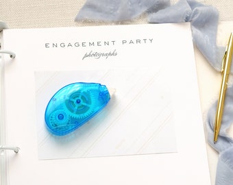 May include: A white binder with the text "ENGAGEMENT PARTY photographs" on the cover. A blue tape dispenser is on the page, and a gold pen and blue ribbon are on the right side of the binder.