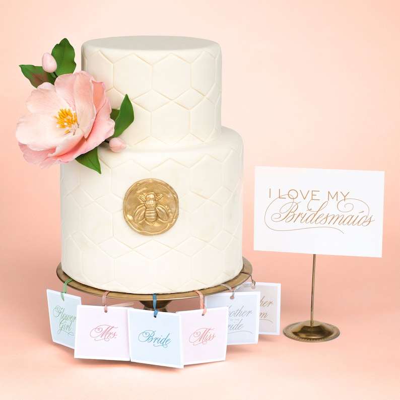 May include: A two-tiered white wedding cake with a honeycomb pattern and a gold bee emblem. The cake is decorated with a pink flower and green leaves.  The cake is on a gold stand with five small tags hanging from it. The tags are labeled "Flower Girl", "Miss", "Bride", "Miss", and "Mother of the Bride". A sign reads "I Love My Bridesmaids".