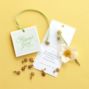 May include: A white card with a green ribbon attached to it. The card has a quote from Winnie the Pooh that reads "The smallest things take up the most room in your heart." The card also has the text "I love you, sweet girl!" written on it. There are several small gold flower charms scattered around the card.