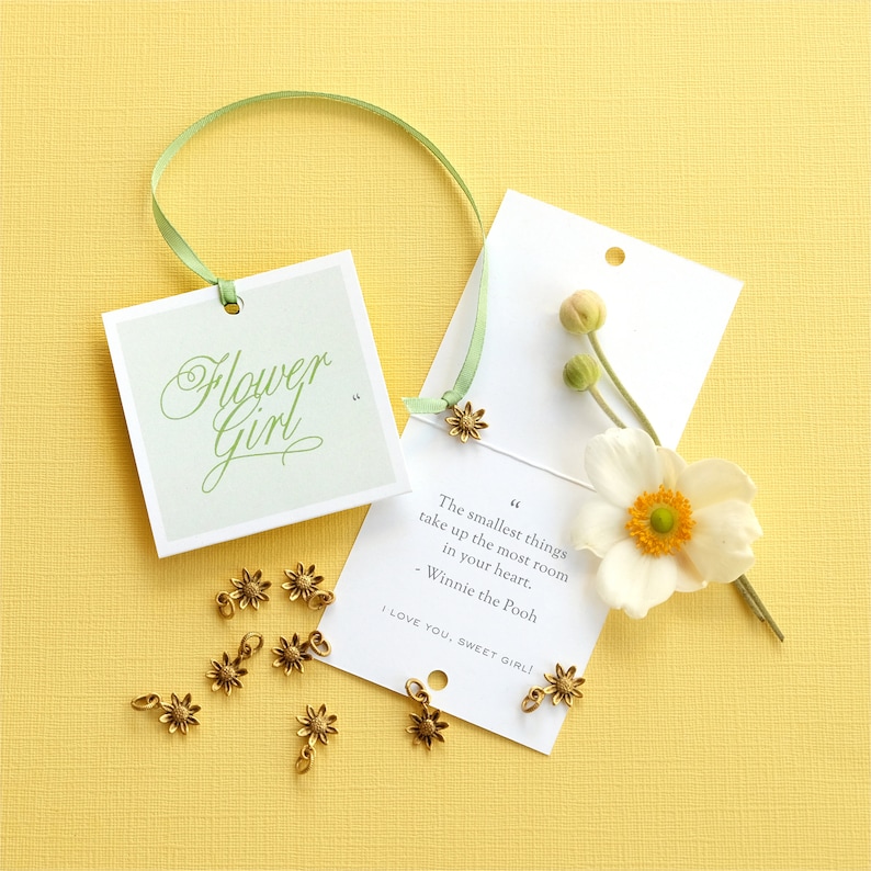 May include: A white card with a green ribbon attached to it. The card has a quote from Winnie the Pooh that reads "The smallest things take up the most room in your heart." The card also has the text "I love you, sweet girl!" written on it. There are several small gold flower charms scattered around the card.