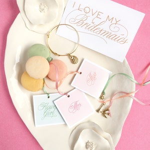 May include: A white ceramic plate with a decorative flower design, featuring a gold bracelet with a charm, four pastel macarons, and four small cards with the words "Miss", "Mrs.", "Flower Girl", and "I Love My Bridesmaids".