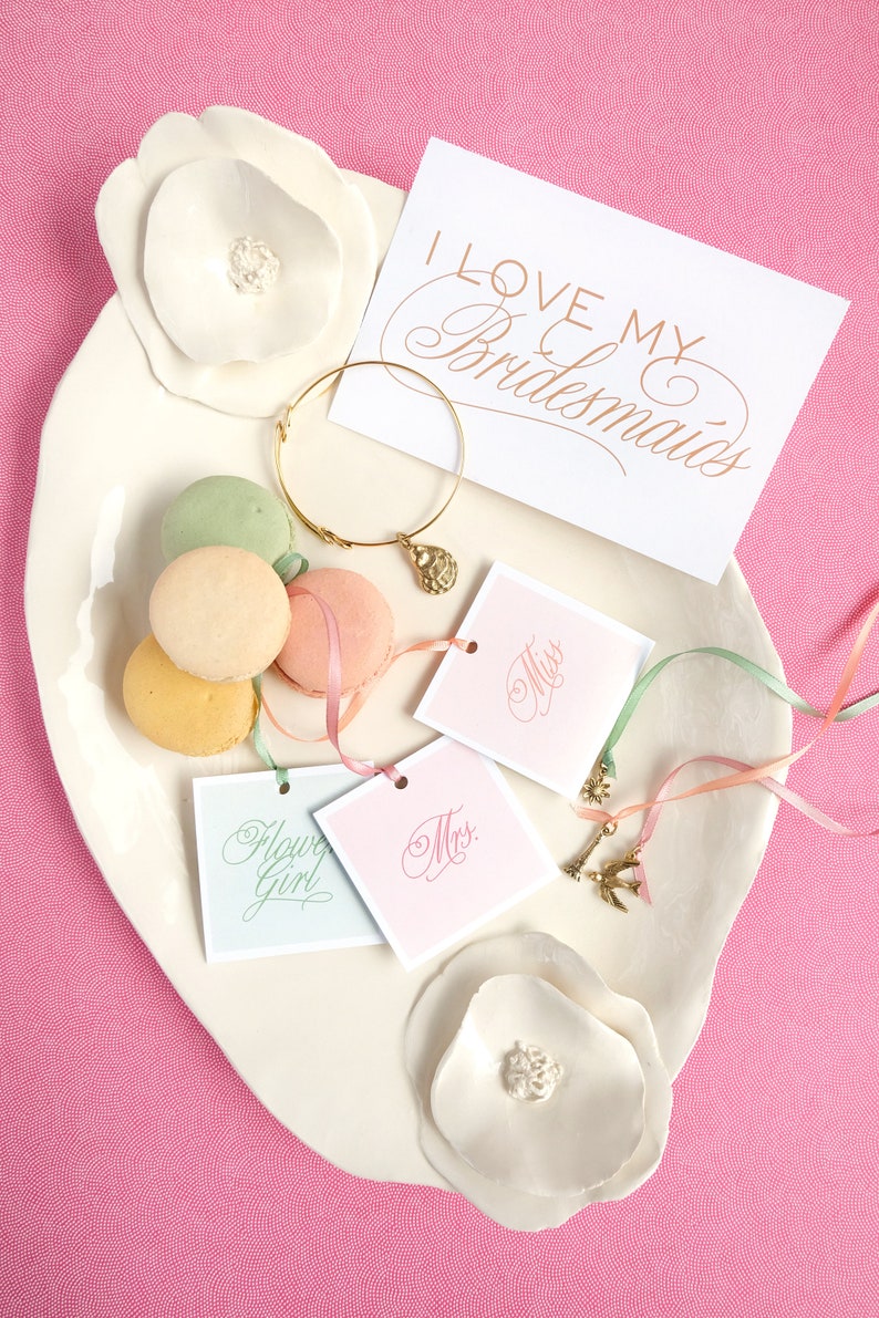 May include: A white ceramic plate with a decorative flower design, featuring a gold bracelet with a charm, four pastel macarons, and four small cards with the words "Miss", "Mrs.", "Flower Girl", and "I Love My Bridesmaids".