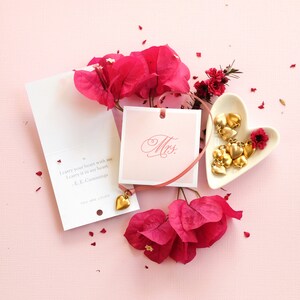 May include: A pink card with the text "I carry your heart with me I carry it in my heart. -E. E. Cummings" and the word "Mrs." on a separate card. The cards are surrounded by pink flowers and a small heart-shaped dish with gold charms.