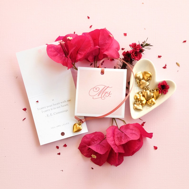 May include: A pink card with the text "I carry your heart with me I carry it in my heart. -E. E. Cummings" and the word "Mrs." on a separate card. The cards are surrounded by pink flowers and a small heart-shaped dish with gold charms.