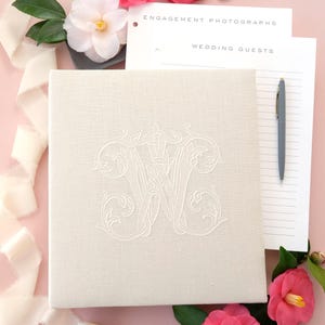 Pode incluir: Um &aacute;lbum de casamento creme com um monograma bordado, rodeado por flores rosa e vegeta&ccedil;&atilde;o. O &aacute;lbum est&aacute; ao lado de papel pautado e uma caneta, com as palavras "Engagement Photographs" e "Wedding Guests" vis&iacute;veis. Uma fita creme tamb&eacute;m est&aacute; presente.