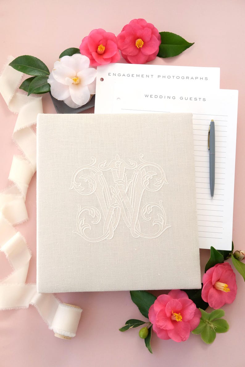 Pode incluir: Um &aacute;lbum de casamento creme com um monograma bordado, rodeado por flores rosa e vegeta&ccedil;&atilde;o. O &aacute;lbum est&aacute; ao lado de papel pautado e uma caneta, com as palavras "Engagement Photographs" e "Wedding Guests" vis&iacute;veis. Uma fita creme tamb&eacute;m est&aacute; presente.