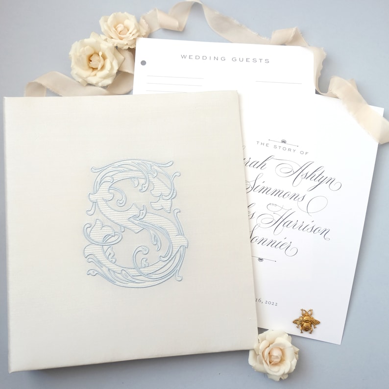 May include: A white wedding guest book with a blue embroidered letter "S" on the front cover. The book is open to reveal a white page with the text "The Story of Sarah Ashlyn Simmons & Harrison Conner" and the date "16, 2022".