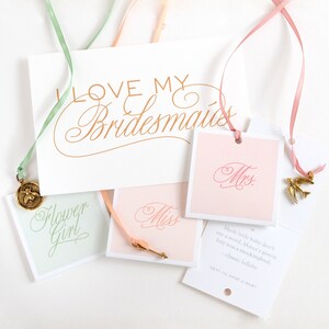 May include: A set of four gift tags with pink, peach, and green ribbons. The tags feature calligraphy text that says "Miss", "Mrs", "Flower Girl", and "Hush little baby don't say a word, Mama's gonna buy you a mockingbird. -classic lullaby NEXT TO HAVE A BABY".