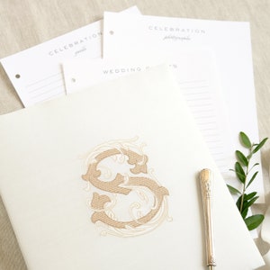 May include: A white wedding guest book with a gold embroidered letter S in a decorative floral design. The book is open to a blank page with a silver pen on top.