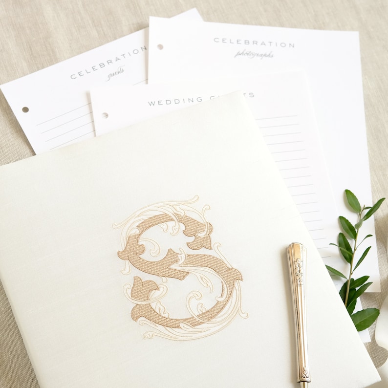 May include: A white wedding guest book with a gold embroidered letter S in a decorative floral design. The book is open to a blank page with a silver pen on top.