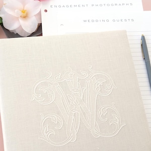 Pode incluir: Livro de visitas de casamento em linho creme com monograma bordado. O livro est&aacute; aberto em p&aacute;ginas pautadas com uma caneta ao lado. Flores rosa e brancas s&atilde;o dispostas perto do livro, com as palavras "Engagement Photographs" e "Wedding Guests" vis&iacute;veis.