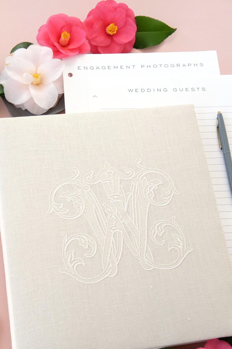 Pode incluir: Livro de visitas de casamento em linho creme com monograma bordado. O livro est&aacute; aberto em p&aacute;ginas pautadas com uma caneta ao lado. Flores rosa e brancas s&atilde;o dispostas perto do livro, com as palavras "Engagement Photographs" e "Wedding Guests" vis&iacute;veis.