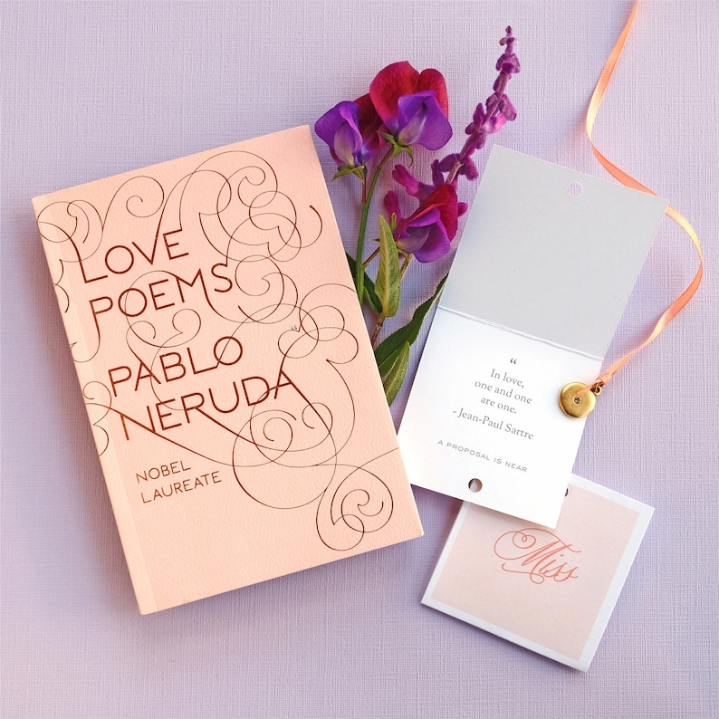 May include: A pink book with the title "Love Poems" by Pablo Neruda, a Nobel Laureate, with a gold-colored design. A white card with the quote "In love, one and one are one." by Jean-Paul Sartre, and the text "A proposal is near." A small white tag with the word "Miss" written in cursive.
