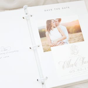 Pode incluir: &Aacute;lbum de casamento branco aberto com uma foto de um casal. O &aacute;lbum inclui um cart&atilde;o "Save the Date" com nomes e uma data, e uma cita&ccedil;&atilde;o: "Falling in love. That is the beginning of everything."