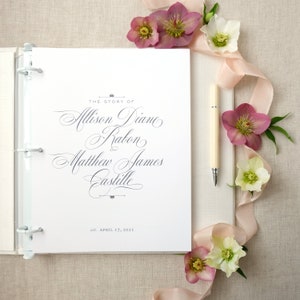 Pode incluir: Um &aacute;lbum de casamento branco com argolas prateadas, aberto numa p&aacute;gina com uma escrita elegante que diz "The Story of Allison Diane Rabon and Matthew James Castille". Uma caneta creme repousa ao lado do &aacute;lbum, com flores rosa e brancas e uma fita.