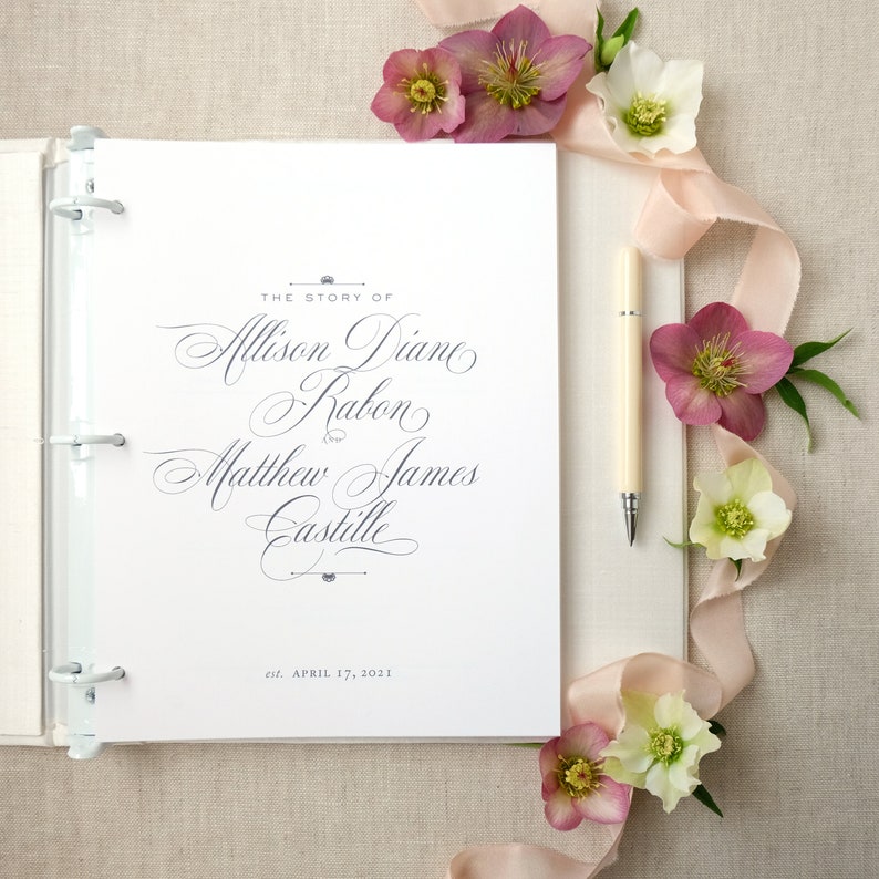 Pode incluir: Um &aacute;lbum de casamento branco com argolas prateadas, aberto numa p&aacute;gina com uma escrita elegante que diz "The Story of Allison Diane Rabon and Matthew James Castille". Uma caneta creme repousa ao lado do &aacute;lbum, com flores rosa e brancas e uma fita.