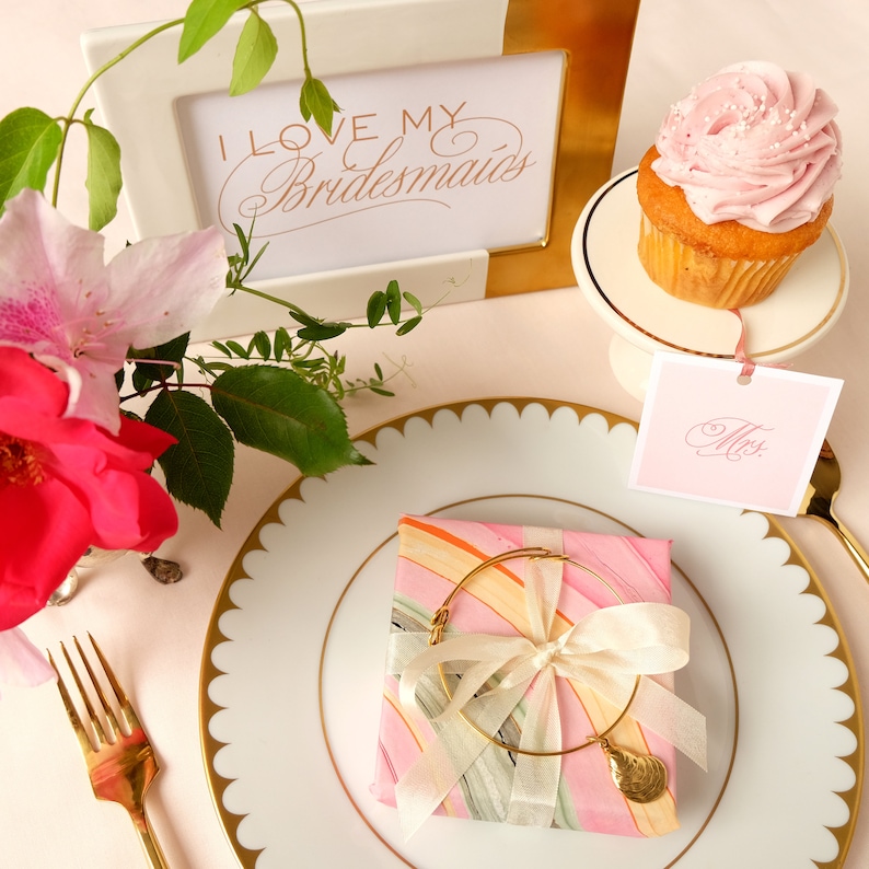 May include: A pink and white marbled gift box with a gold bracelet and a pink and white ribbon. The box is on a white plate with a gold rim. The plate is on a pink table with a cupcake and a sign that says "Mrs."