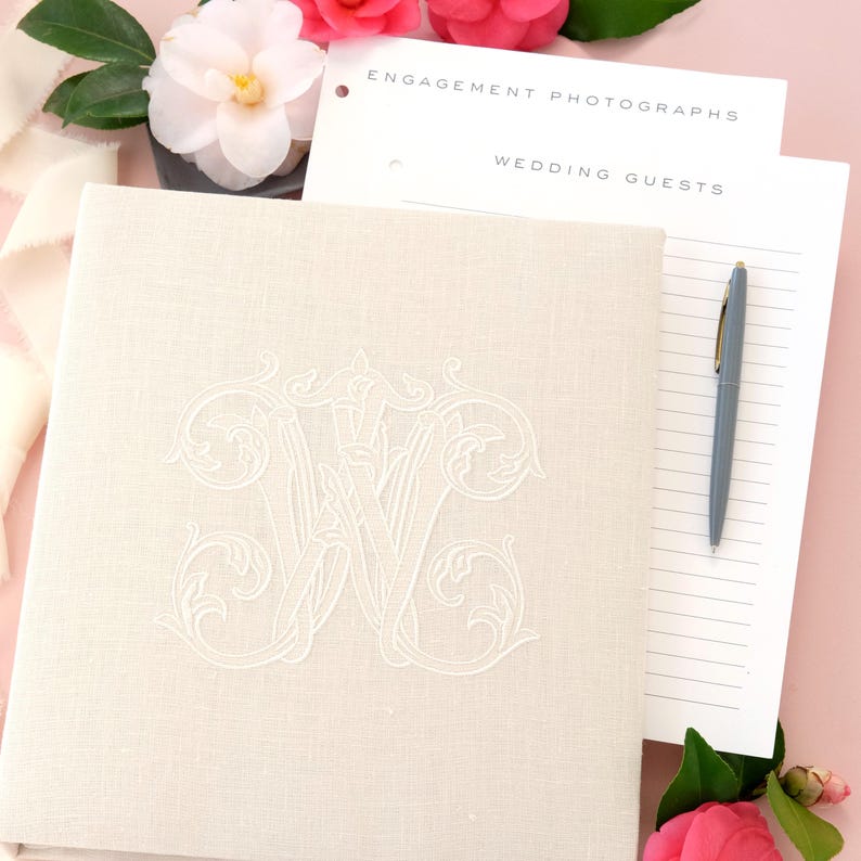 May include: A white linen wedding album with a white embroidered monogram of the letters 'JW' in a decorative font. The album is lying on a pink surface with a white notepad with the words 'Engagement Photographs' and 'Wedding Guests' written on it. A gray pen is lying on the notepad.