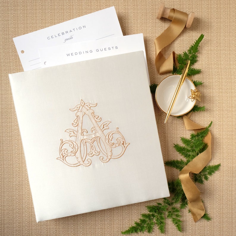 May include: A white wedding guest book with a gold embossed letter A on the front. The book is lying on a brown surface with a gold ribbon and green foliage.