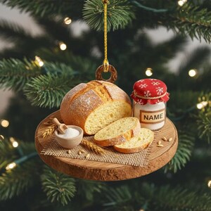May include: A round, wooden Christmas ornament featuring a loaf of bread, sliced bread, a small bowl of flour, and a jar with the text "NAME". The ornament is decorated with wheat stalks and a red and white lid.