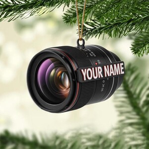 May include: A black camera lens Christmas ornament with a red accent ring and the text "YOUR NAME" in white. The ornament is hanging from a gold string against a blurred background of green pine needles.