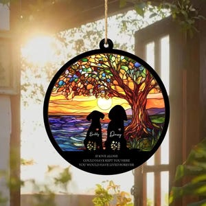 May include: A round, black decorative hanging ornament featuring a stained-glass design. The image depicts two dog silhouettes under a tree at sunset, with the names "Buddy" and "Daisy" and the quote "If love alone could have kept you here, you would have lived forever."