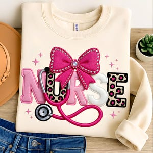May include: Cream-colored sweatshirt with the word "NURSE" in pink and white, featuring a pink bow with rhinestones and a stethoscope. The letter "E" has a leopard print design. Pink stars are embroidered around the text.