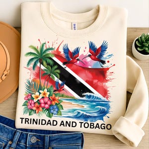 May include: Cream-colored sweatshirt featuring a vibrant graphic of Trinidad and Tobago. The design includes the national flag, palm trees, tropical flowers, and scarlet ibis birds. The text "TRINIDAD AND TOBAGO" is printed at the bottom.